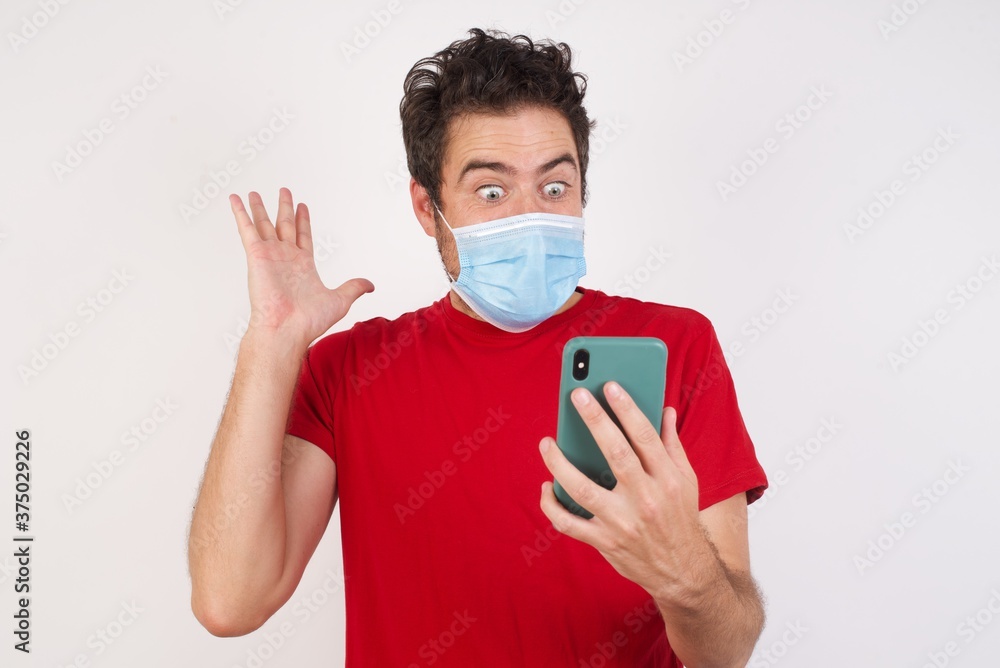 Close-up portrait of Young caucasian man with short hair wearing medical mask standing over isolated white background holding in hands cell reading browsing news Close-up portrait of Young caucasian man with short hair wearing medical mask standing over isolated white background holding in hands cell reading browsing news