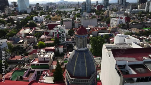 Mixcoac church tower, steeple, dome, and steel cross in downtown Mexico city center buildings, red roofs and skyline, rising tilt down circle aerial photo