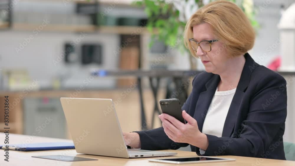 Old Businesswoman Working on Laptop and Smartphone in Office Old Businesswoman Working on Laptop and Smartphone in Office