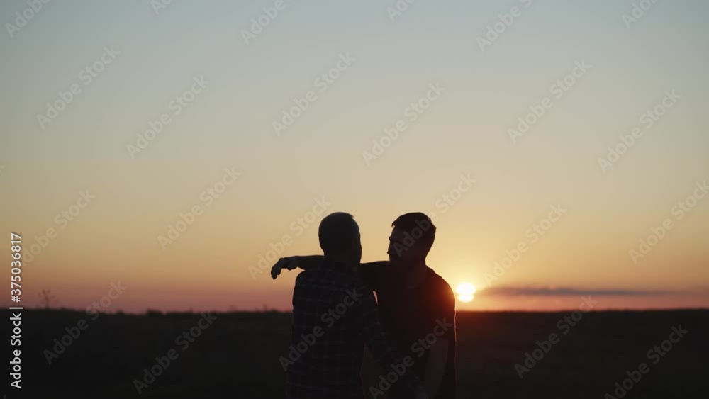 Smiling young attractive man son embracing his old father at sunset in summer twilight. Enjoyment of family ties. Trust and love between generations. Human aging. Scenic landscape on background. 4K Smiling young attractive man son embracing his old father at sunset in summer twilight. Enjoyment of family ties. Trust and love between generations. Human aging. Scenic landscape on background. 4K