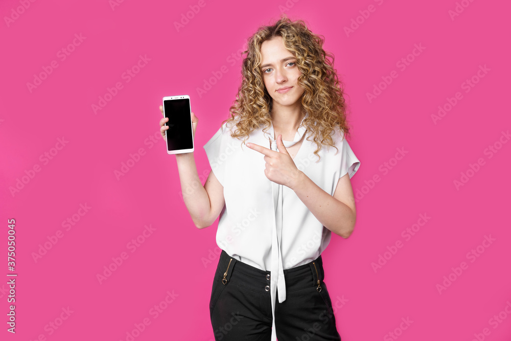 Look at this cell phone. Contented happy woman, pointing her index finger at a blank screen, shows a modern device. Isolated on a pink background. Look at this cell phone. Contented happy woman, pointing her index finger at a blank screen, shows a modern device. Isolated on a pink background.