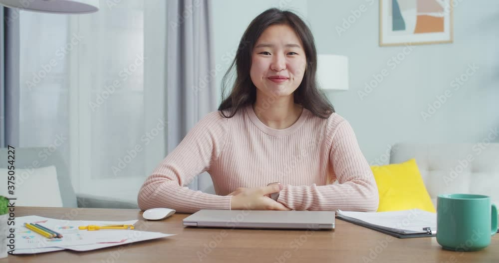 Confident adult Asian woman sitting at table at home office room. Beautiful female student with dimples in casual clothes looking at camera and smiling. Modern minimalist design. Confident adult Asian woman sitting at table at home office room. Beautiful female student with dimples in casual clothes looking at camera and smiling. Modern minimalist design.