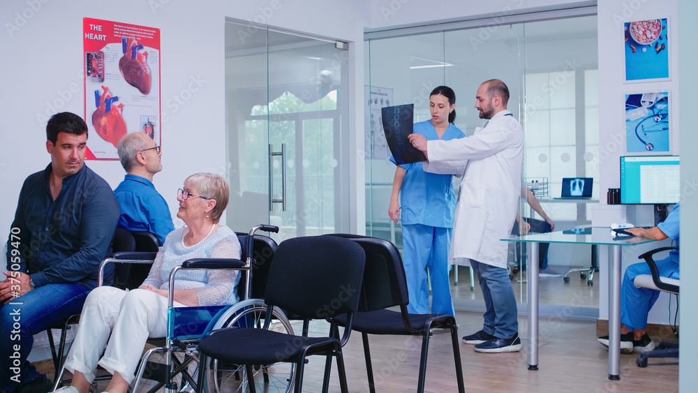 Fototapeta premium Doctor holding patient x-ray explaining diagnosis to nurse in waiting area. Disabled senior woman wheelchair waiting for medical examination. Hospital and health care system, medicine private clinic