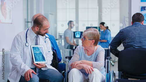 Doctor holding tablet pc with x-ray while explaining diagnosis to disabled elderly woman in wheelchair. Handicapped patient in hospital waiting area. Man in examination room.