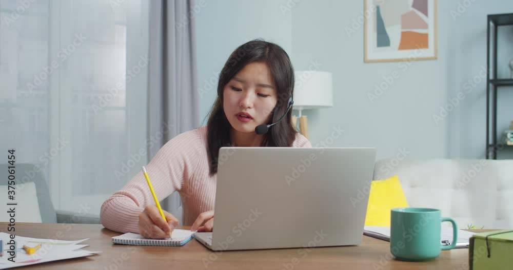 Attractive adult female worker in headphones sitting at table and talking to client, customer. Young Asian woman working at home office and taking notes. Modern minimalist interior design. Attractive adult female worker in headphones sitting at table and talking to client, customer. Young Asian woman working at home office and taking notes. Modern minimalist interior design.