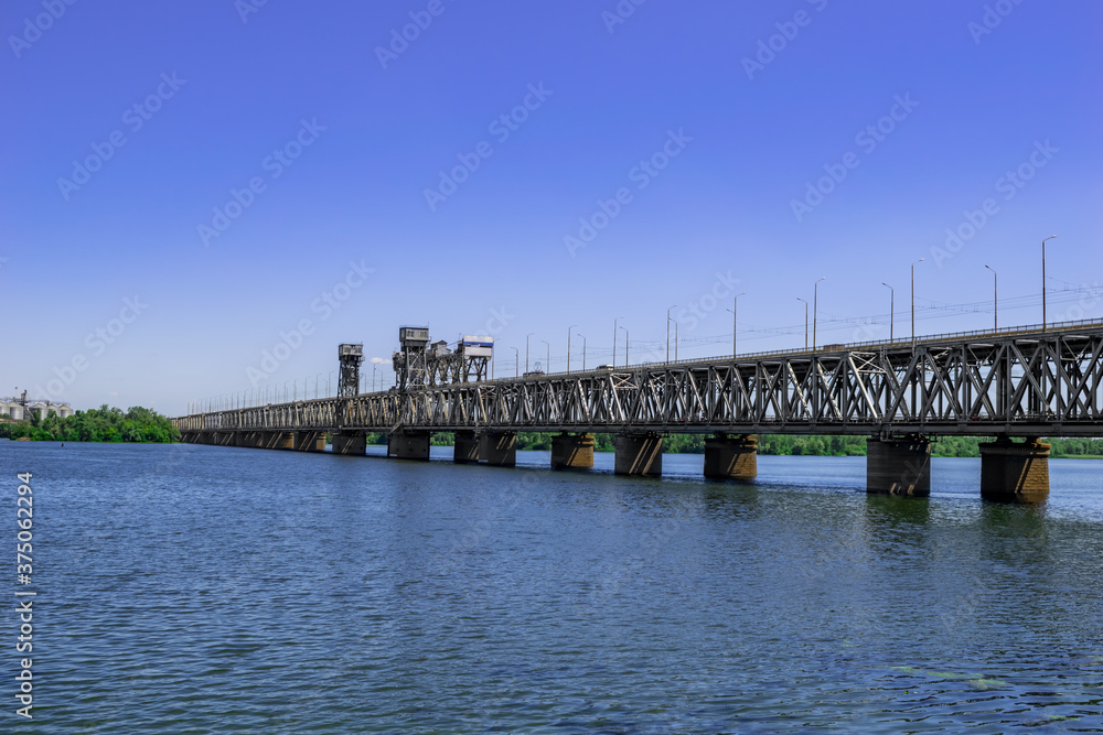 Metal Amur bridge over the Dnieper river in Dnipro (Ukraine), side view. Modern monument of engineering art against the background of a blue water surface. Background with copy space