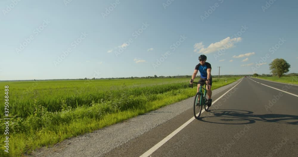 A cyclist is riding on the road. Sportsman is training on a bicycle. Green field and the sun is shining in the background. 4K A cyclist is riding on the road. Sportsman is training on a bicycle. Green field and the sun is shining in the background. 4K