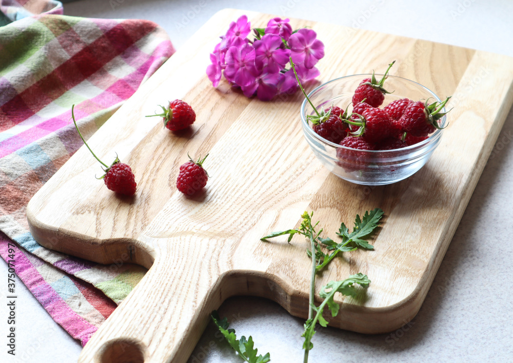 Ripe raspberries in a dish and scattered on a wooden Board with flowers and a background of a checkered napkin, side view-the concept of using vitamin berries in cooking