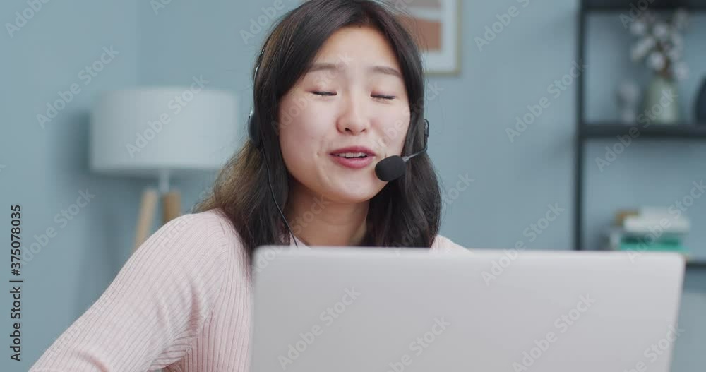 Beautiful Asian woman in headphones sitting at table and using laptop. Young female student talking to famiy, clients, friend via video webcam chat. Relationship, friendship, social media. Beautiful Asian woman in headphones sitting at table and using laptop. Young female student talking to famiy, clients, friend via video webcam chat. Relationship, friendship, social media.