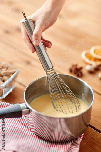 cooking, culinary and seasonal drinks concept - close up of hand with whisk stirring eggnog in pot