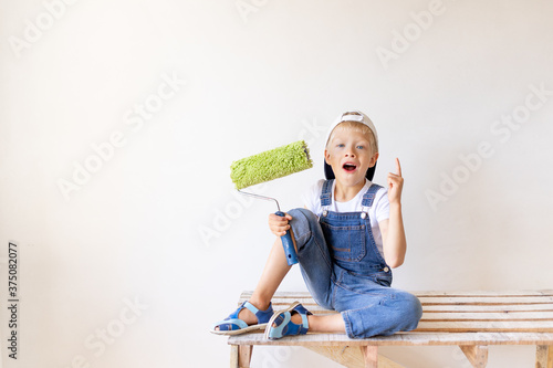 a surprised child Builder sits on a construction ladder in an apartment with white walls and a roller in his hands and shows a thumbs up, place for text, repair concept