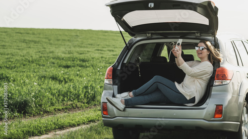Young Woman Sitting in Open Car Trunk and Using Smartphone for Video Call © Romvy