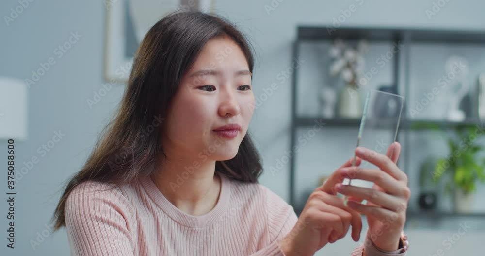 Portrait of young Asian woman using clear glass smart phone at home. Pretty female student scrolling news feed, chatting, watching video, photos. Futuristic cyber technology concept. Portrait of young Asian woman using clear glass smart phone at home. Pretty female student scrolling news feed, chatting, watching video, photos. Futuristic cyber technology concept.