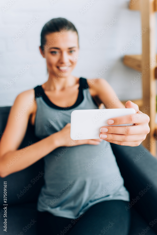 Selective focus of pregnant sportswoman taking selfie with smartphone on sofa at home
