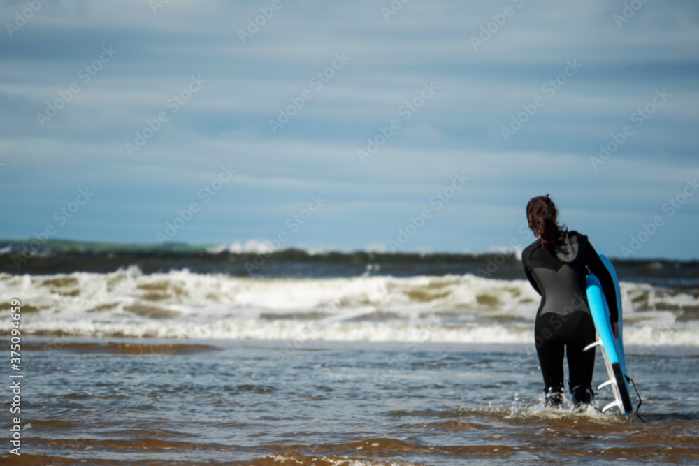 Fit female surfer in black swimsuit walking into ocean with her surf board, Blue cloudy sky, Selective focus. Concept outdoor activity, sport and healthy lifestyle. Fit female surfer in black swimsuit walking into ocean with her surf board, Blue cloudy sky, Selective focus. Concept outdoor activity, sport and healthy lifestyle.