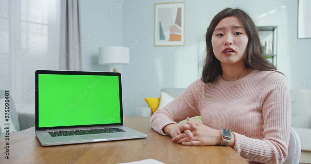 Young Asian woman sitting at table with chroma key, green screen on laptop computer. Beautiful female representative speaking, explaining on camera and gesturing at home. Commerce, business. Young Asian woman sitting at table with chroma key, green screen on laptop computer. Beautiful female representative speaking, explaining on camera and gesturing at home. Commerce, business.