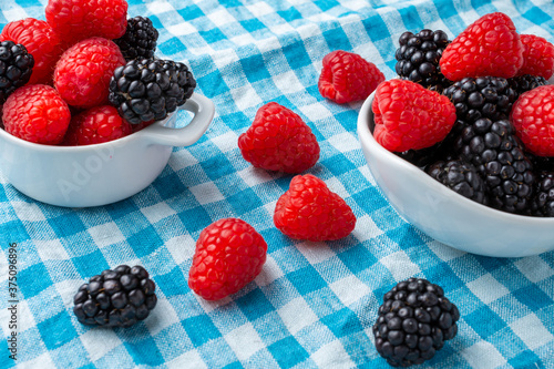 Top view of two white bowls with blackberries and raspberries  on blue checkered cloth  with fruits  horizontal