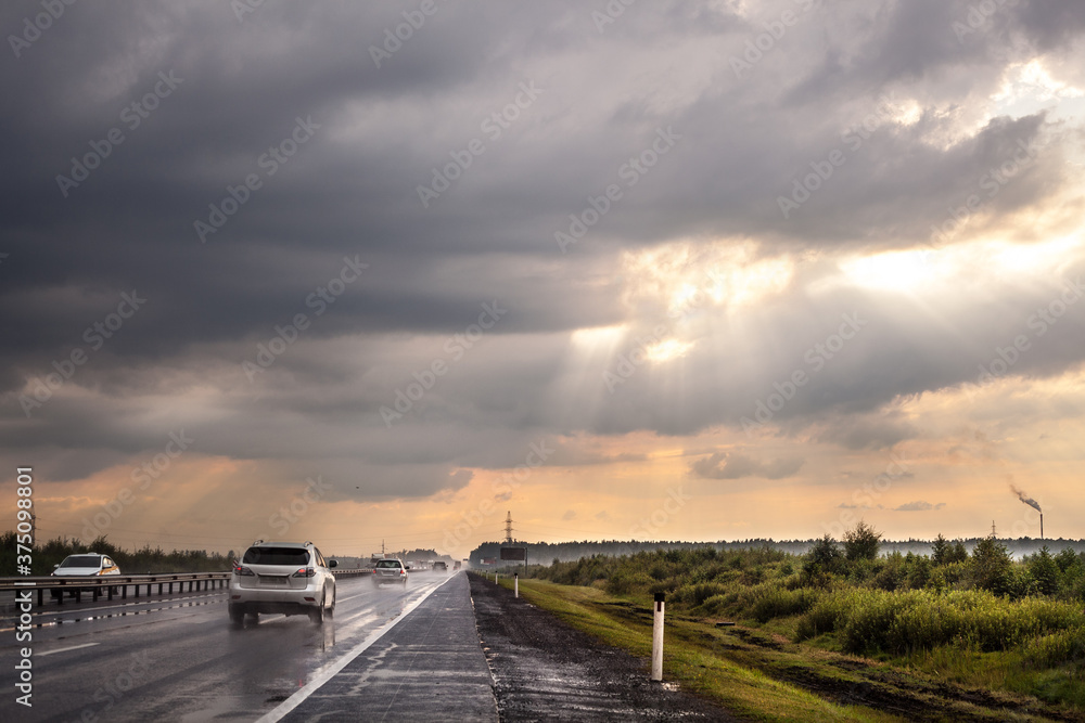 Fototapeta premium Highway after rain. Cars are driving along the road. Grass, trees and bushes grow on the side of the road. Cloudy sky. The sun's rays make their way through the clouds. Fog in the distance.