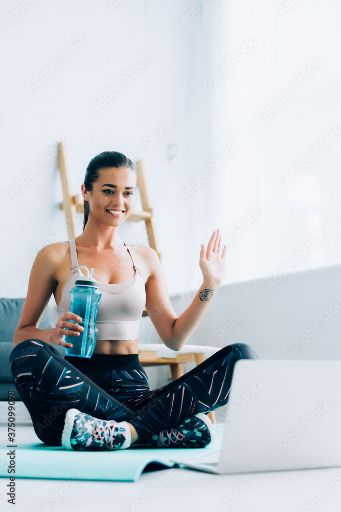 Selective focus of sportswoman waving hand while holding sports bottle near laptop at home Selective focus of sportswoman waving hand while holding sports bottle near laptop at home