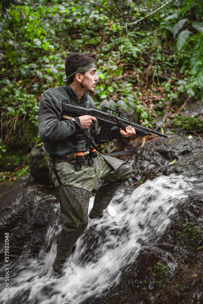 Soldier or revolutionary member or hunter in camouflage in the stream observing the gun in his hand