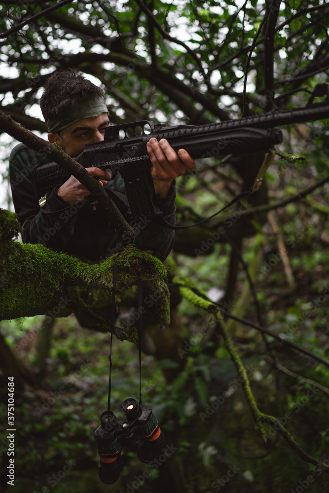 Soldier or revolutionary member or hunter in camouflage on the tree aiming the gun in his hand, hunter concept
