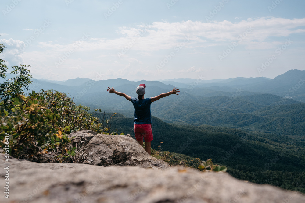 Beautiful rocky mountainous terrain and mountain peaks with dense forest in the distance. A young female tourist stands on top of a mountain with her arms spread wide as if embracing the whole world.