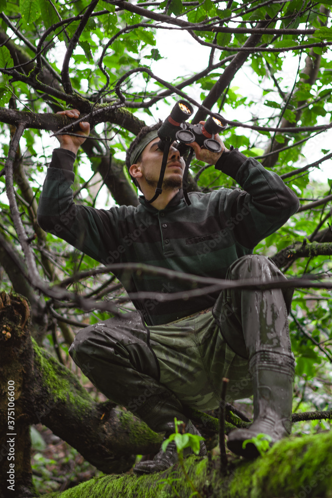 Soldier or revolutionary member or hunter in camouflage on the tree observing the binocular in his hand