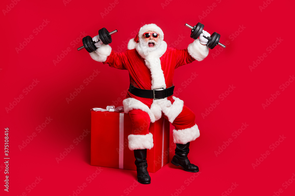 Full length photo of retired old man white beard sit giftbox hold dumbbells crazy strength scream wear x-mas santa costume glove coat belt sunglass cap boot isolated red color background Full length photo of retired old man white beard sit giftbox hold dumbbells crazy strength scream wear x-mas santa costume glove coat belt sunglass cap boot isolated red color background