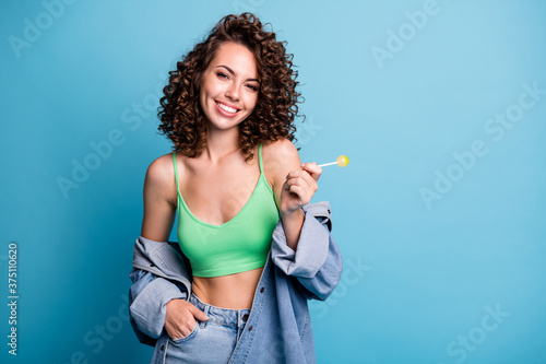 Photo of lovely attractive nice girl youngster enjoy summer dream weekend hold tasty delicious yummy lollipop wear green denim clothes isolated over blue color background