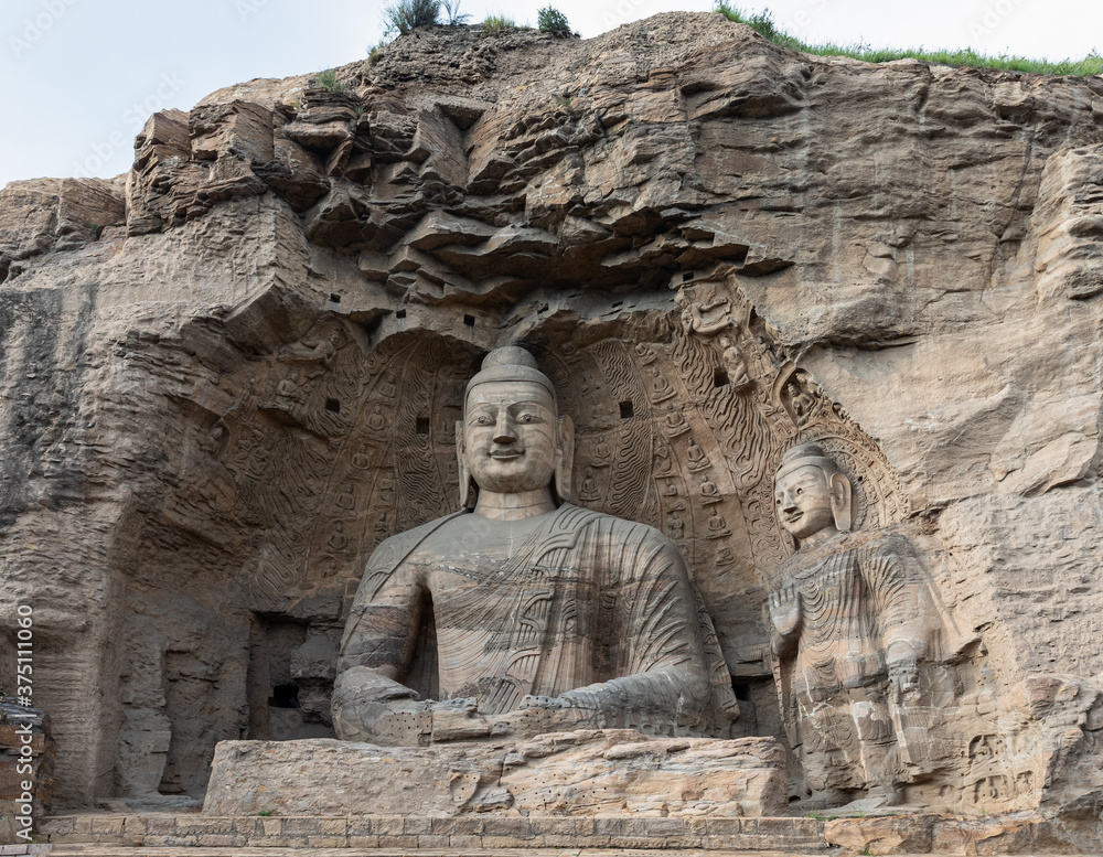 Iconic seated Buddha statue and smaller standing Buddha on right in Cave 20 at Yungang Grottoes, Datong, Shanxi, China. Constructed from 5th century. UNESCO World Heritage.