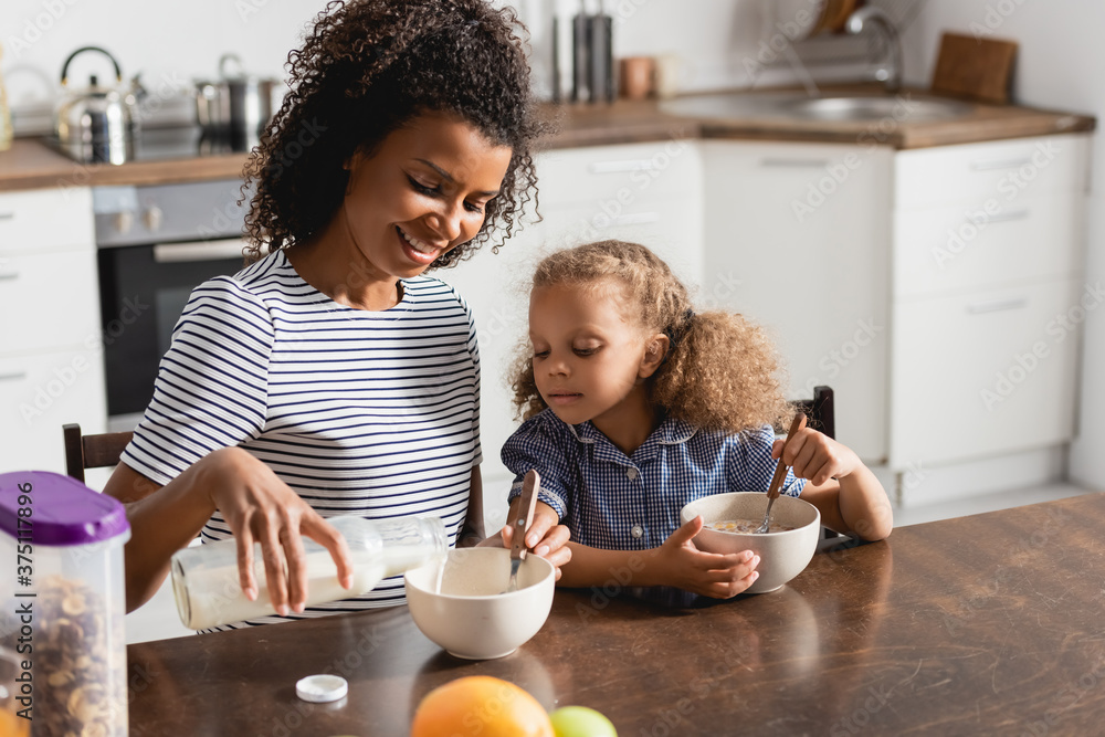 african american mom in striped t-shirt pouring milk into bowl while having breakfast with daughter