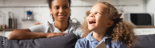 selective focus of young african american nanny behind excited girl laughing while looking away, horizontal image photo