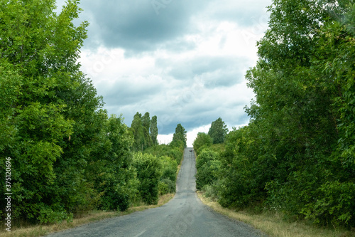 Beautiful sceneric view at the road among green trees and brushes, cloud sky. The road goes up to the sky.
