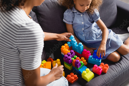 cropped view of babysitter near african american girl in dress playing with multicolored building blocks