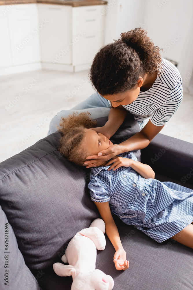 Fototapeta premium high angle view of african american woman touching face of daughter sitting on couch in dress near toy bunny