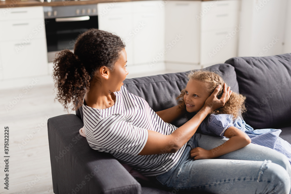 young african american mother touching head of excited daughter while sitting on sofa at home young african american mother touching head of excited daughter while sitting on sofa at home