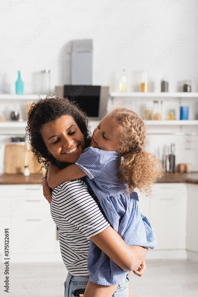 Obraz premium young african american woman with closed eyes holding daughter on hands in kitchen