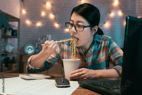 elegant businesswoman eating instant noodles at dining table while over work on project with laptop computer and calculator. hungry asian korean female in glasses having bedtime snacks in night.