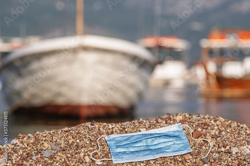 A discarded medical face mask contaminates a beautiful sandy pebble beach, with fishing boats in the background. Budva, Montenegro