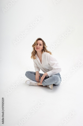 portrait of young smiling caucasian woman posing in shirt and blue jeans, sitting on white studio floor. model tests of pretty girl in basic clothes on cyclorama. attractive female poses, smiles