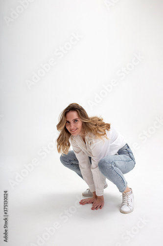portrait of young smiling caucasian woman posing in shirt and blue jeans, sitting on white studio floor. model tests of pretty girl in casual clothes on cyclorama. attractive female fooling around