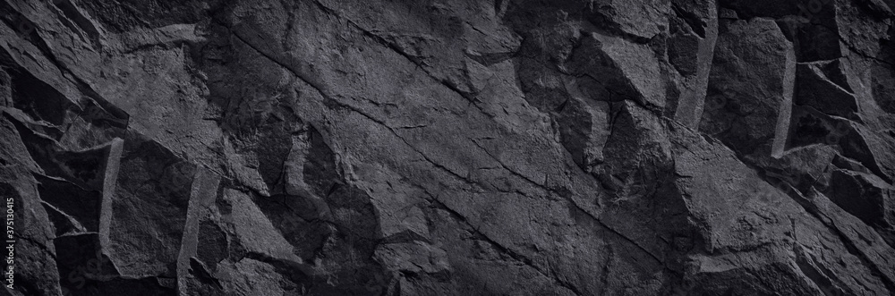 Fototapeta Czarny kamień tło. Ciemna tekstura skały. Tekstura powierzchni górskiej. Zbliżenie. Szeroki baner o wolumetrycznej fakturze kamienia. Czarne tło z miejsca kopiowania dla swojego projektu.