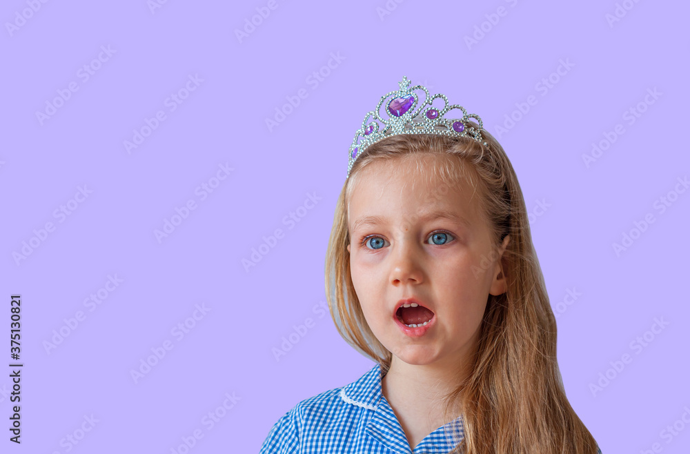 Surprised little princess girl in silver crown looking away with open mouth. Open eyed emotional young lady with long wavy hair in blue dress portrait on pastel purple background with copy space. Surprised little princess girl in silver crown looking away with open mouth. Open eyed emotional young lady with long wavy hair in blue dress portrait on pastel purple background with copy space.