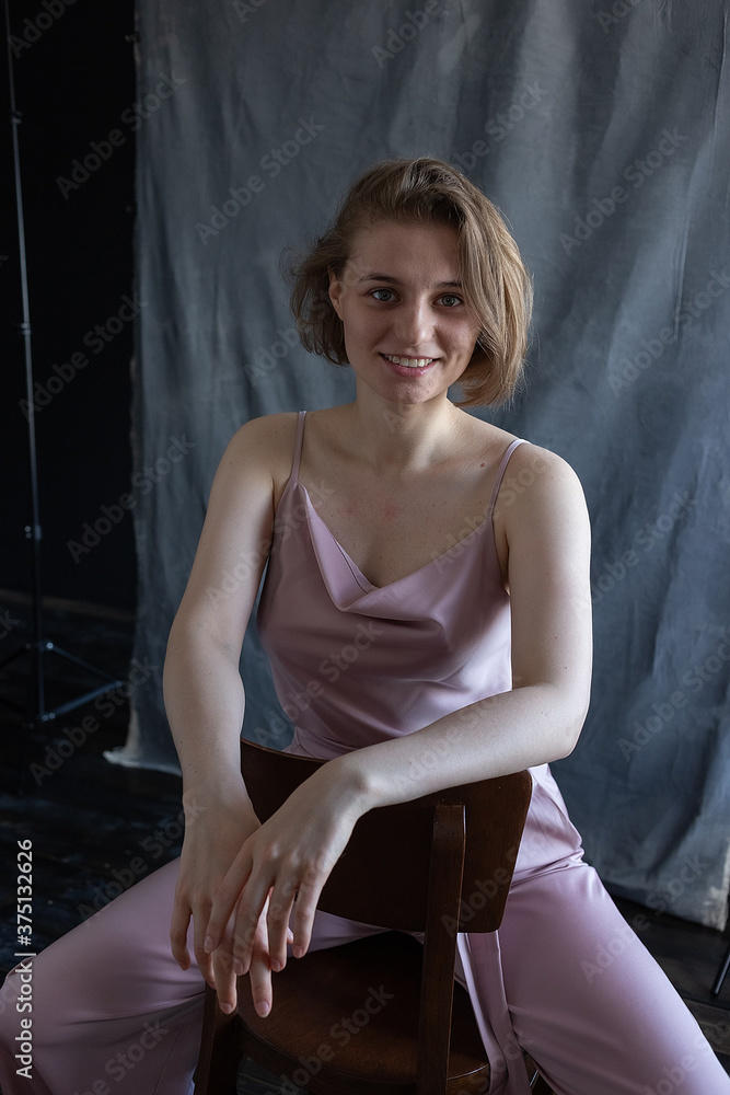 portrait of young smiling caucasian woman with short hair posing in pink suit, sitting on chair in front of fabric background. pretty girl in blouse. attractive female poses and smiles portrait of young smiling caucasian woman with short hair posing in pink suit, sitting on chair in front of fabric background. pretty girl in blouse. attractive female poses and smiles