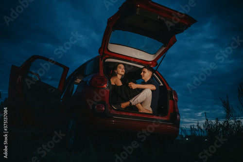 Couple in love sits and holds each other hands inside of opened trunk of car at night.
