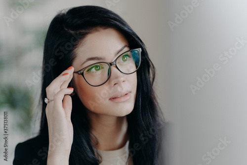 Close up shot of brunette serious lady keeps hand on rim of spectacles, concentrated at computer screen, watches online video, poses indoor, browses web page, creats project, poses in office