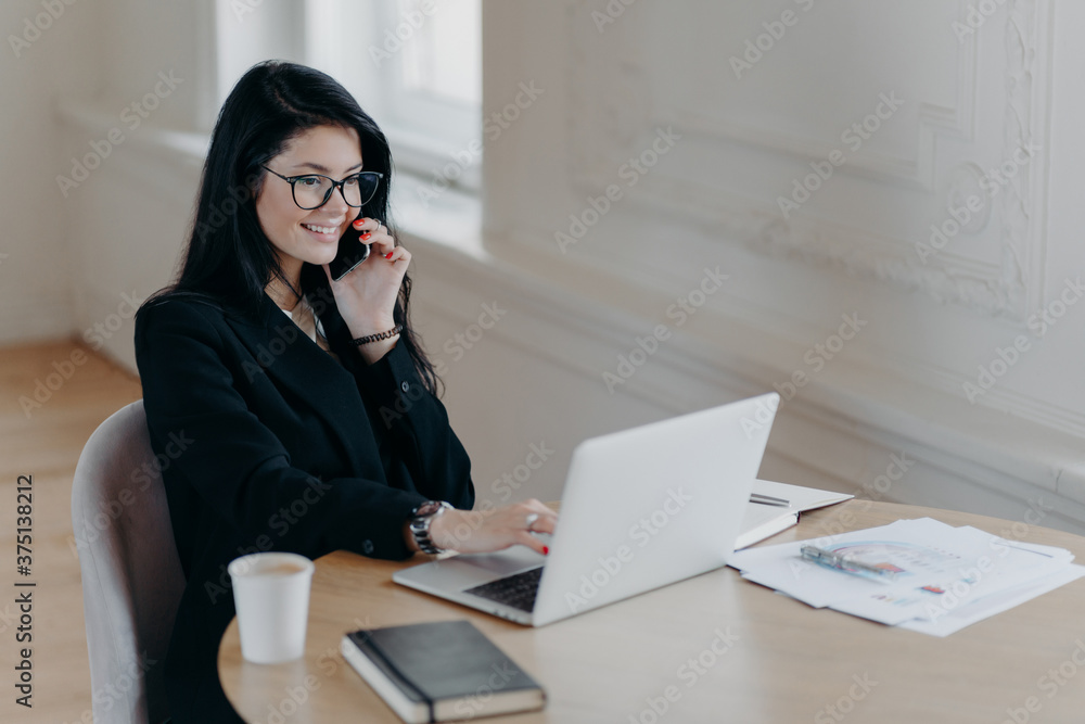 Beautiful female executive makes remote job, talks via mobile phone and checks information in laptop computer poses at workplace surrounded by coffee paper documents notepad has telephone conversation Beautiful female executive makes remote job, talks via mobile phone and checks information in laptop computer poses at workplace surrounded by coffee paper documents notepad has telephone conversation
