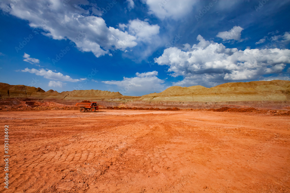 Bauxite clay quarry. Aluminium ore mining and transporting. Open-cut mine and orange Hitachi dump truck. On blue sky with white clouds background.