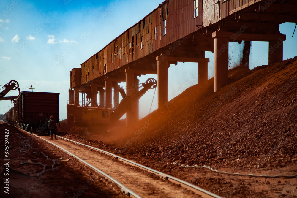 Obraz premium Bauxite clay open-cut mining. Loading and railway transporting terminal. Excavator load aluminium ore. Hopper car train on rails. Worker controlling process.