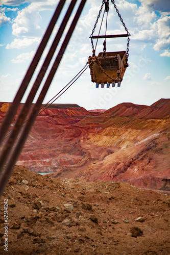 Aluminium ore quarry. Red bauxite clay open-cut mining. Walking dragline excavator bucket on cable of mast at work. Blue sky, clouds.
